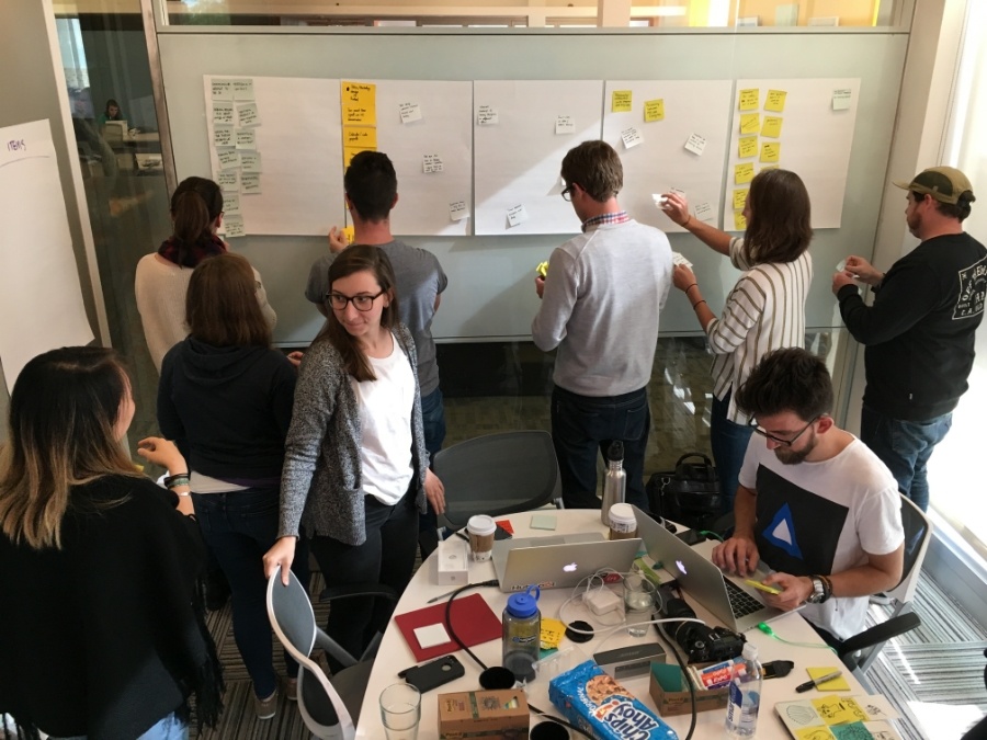 Photograph from a Hubspot Design Thinking Workshop. There are 9 people in a small room, 5 are sticking sticky notes on a whiteboard, and the others are standing or sitting around a small circular table.